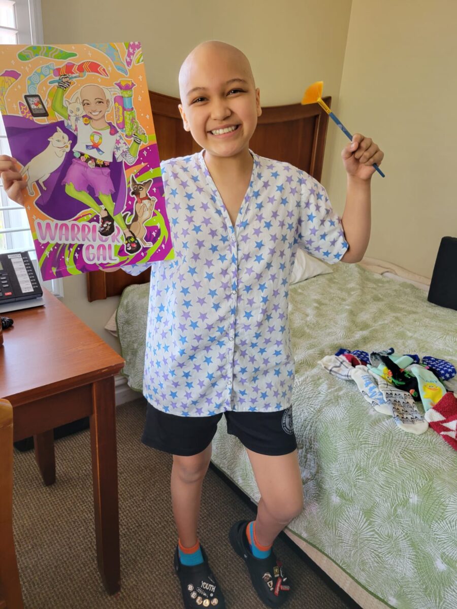 Photo of a smiling bald girl who is holding up a poster of her superhero self.
